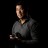 Pixelated static image of a person in profile view wearing a dark jacket over a light shirt against a black background.