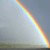 A diagonal rainbow arc spans from bottom-right to top-left against a muted gray background with faint greenish terrain at the bottom.