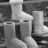 Grayscale pixelated image of a stylized figure with light-colored hair, wearing a long dress and boots, standing against a dark background.