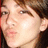 Pixelated close-up of a person's face with light skin, brown hair, and visible eyes, nose, mouth, and ear against dark background.