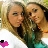 Two people in close proximity, one with light brown hair and the other with dark hair, both wearing dark tops with green accents, against a blurred background with a small pink and black pixelated shape in the bottom left.