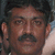 A static image of a person with dark hair and a mustache, wearing a light-colored shirt, against a blurred background.