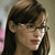 A pixelated close-up of a person with dark hair and glasses, wearing a dark top against a blurred background with pink and beige tones.