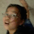 Pixelated close-up of a person with dark hair and glasses, mouth open in laughter against a muted background.