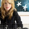 A person with long blonde hair and red lipstick wears a black jacket with white stripes on the sleeves, positioned against a grid-patterned background with two white star shapes.