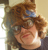 Pixelated static image of a person with brown curly hair and light skin, cropped to show upper face and shoulders against a neutral background.