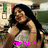 A woman with long dark hair in a light-colored sleeveless top poses against a blurred background, her hand near her neck.