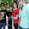 Four people stand outdoors under green trees, wearing casual clothing with visible graphic designs on shirts.
