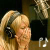 A person with blonde hair wearing large black headphones, positioned near a microphone with musical notes visible in the foreground.