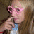 Pixelated image of a person with light brown hair wearing pink heart-shaped glasses and a blue top, holding a dark object near their face.