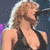A six- shows a woman with long blonde hair singing into a microphone, wearing a black sleeveless top against dark stage lighting