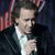 Pixelated image of a person in a dark suit holding a microphone, with red and blue striped background elements.