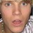 Static pixelated close-up of a person's face with light skin, brown eyes, and parted lips showing teeth, wearing a checkered shirt.