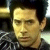 Pixelated close-up of a person with dark hair and light skin wearing a dark patterned shirt against a blurred background.