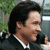 Pixelated static image of a person with dark hair wearing a black suit and white shirt, set against an outdoor background with green foliage and gray structures.