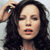 A pixelated close-up of a person with long dark hair, hand raised near head, closed eyes, and mouth slightly open against a light background.