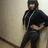 A person in a black hoodie and dark pants stands against a light wall with one hand on their hip, legs crossed.