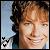 Pixelated portrait of a person with light brown hair and closed eyes, smiling against a blue sky background with black heart symbols.
