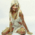 A blonde woman in a white dress sits on the ground with legs spread, smiling at the camera against a light background.
