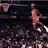 A basketball player in mid-air dribbling a ball, viewed from a low angle with a blurred crowd in the background.