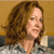 Pixelated image of a person with brown curly hair, light skin, and a neutral expression, wearing a light-colored top against a brown background.