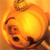 A pixelated orange-yellow circular icon with a dark curved line on the left side and two dark oval shapes below it, set against a warm-toned background.