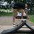 A person in a white tank top and black shorts stands on a seesaw in a park, with trees and grass in the background.