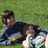 Two people lie on grass; one wears a striped shirt and holds a white object while the other rests their head on the first person's chest.