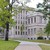 Pixelated static image of a large white building with columns, surrounded by green trees and grass, viewed from a paved path.