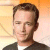 Pixelated portrait of a person with short brown hair wearing a dark jacket over a white shirt against a gradient background of orange and beige tones.
