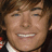 A pixelated close-up of a smiling person with brown hair and light skin, wearing a white collar and dark clothing against a dark background.