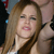 A pixelated close-up of a person with long light brown hair, wearing a dark top with a white strap visible on the shoulder.