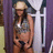 A pixelated image of a person wearing a light-colored cowboy hat, sleeveless top, and dark shorts standing near a white structure with vertical bars.