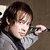 A person with brown hair wearing a gray jacket holds a black object near their face against a brown background.