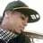 Pixelated image of a person wearing a dark green wide-brimmed hat and a checkered shirt, facing left with a neutral expression.