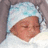 Pixelated image of a person with closed eyes and a neutral expression, wearing a light blue head covering against a gray background.