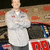 A person in a gray jacket with arms crossed stands before a blurred background with red and blue elements.