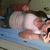 Pixelated image of a person with light skin and brown hair lying on a blue surface, wearing a white top, against a muted green background.