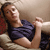 A person in a dark blue sleeveless top reclines on a light-colored couch with a red patterned cushion, holding a white object near their face.