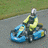 A person in a yellow jacket and silver helmet drives a blue go-kart on a paved track with grassy background.
