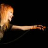 A person with long orange hair extends their right arm forward against a black background, wearing a dark top with a visible microphone cable.