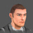 Pixelated portrait of a person with short brown hair, light skin tone, and neutral expression wearing dark jacket over white shirt against gray background.