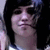 Pixelated close-up of a person with dark hair and light skin, wearing a light-colored top with dark accents, against a dark background.