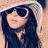 Pixelated image of a person with long dark hair wearing sunglasses and a light-colored hat, facing left against a blurred background.