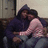 Two figures embrace on a couch; one wears a dark hooded garment and jeans, the other a light pink sleeveless top.