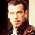 Static pixelated image of a person with dark hair and light skin wearing a dark jacket over a white shirt, set against a gradient background transitioning from black to beige.