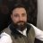 Pixelated static image of a person with light skin, brown hair, and a beard wearing a white shirt under a dark vest against a dark background.