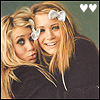 Two blonde women in dark clothing pose closely together, one with a white bow on her head and both with green eyes, against a muted green background with two white heart icons in the top right.