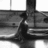Static grayscale image of a stylized figure with dark hair and clothing standing in front of horizontal lines suggesting a background structure.