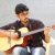 Pixelated image of a person wearing a dark cap and dark shirt playing a light-colored guitar against a plain background.